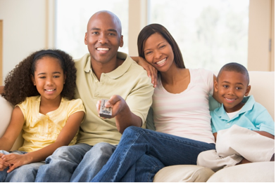 Photo of a girl, a young man, a young woman, and a boy sitting together on a couch. The man is holding a remote control selector.